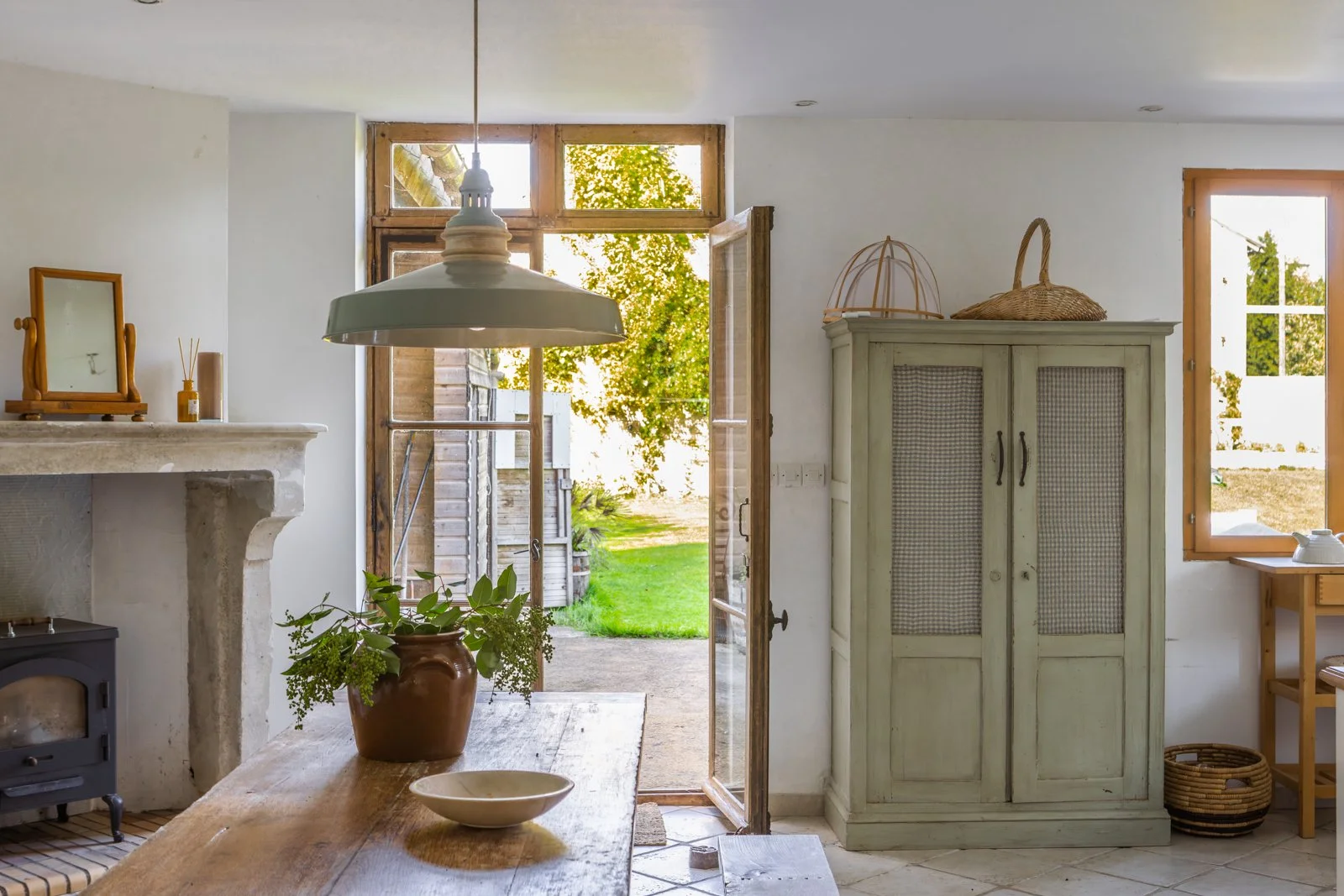 Interior of a rustic kitchen with a wooden table, potted plant, white ceramic bowl, fireplace with a mirror and candles, green cabinet, open door leading outside to a garden, and a window with sunlight streaming in.