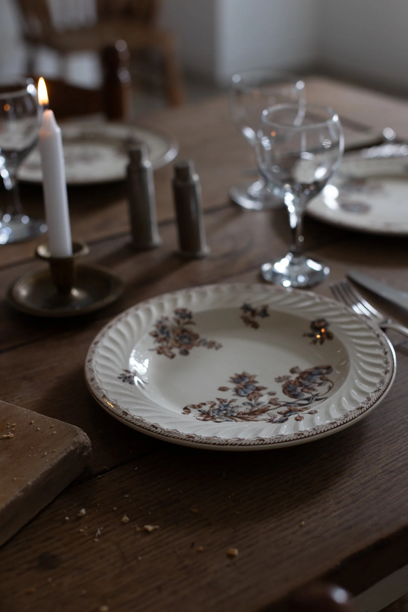 A vintage floral ceramic plate on a wooden dining table set for a meal with wine glasses, a lit candle, and salt and pepper shakers. The table has a rustic, cozy ambiance.