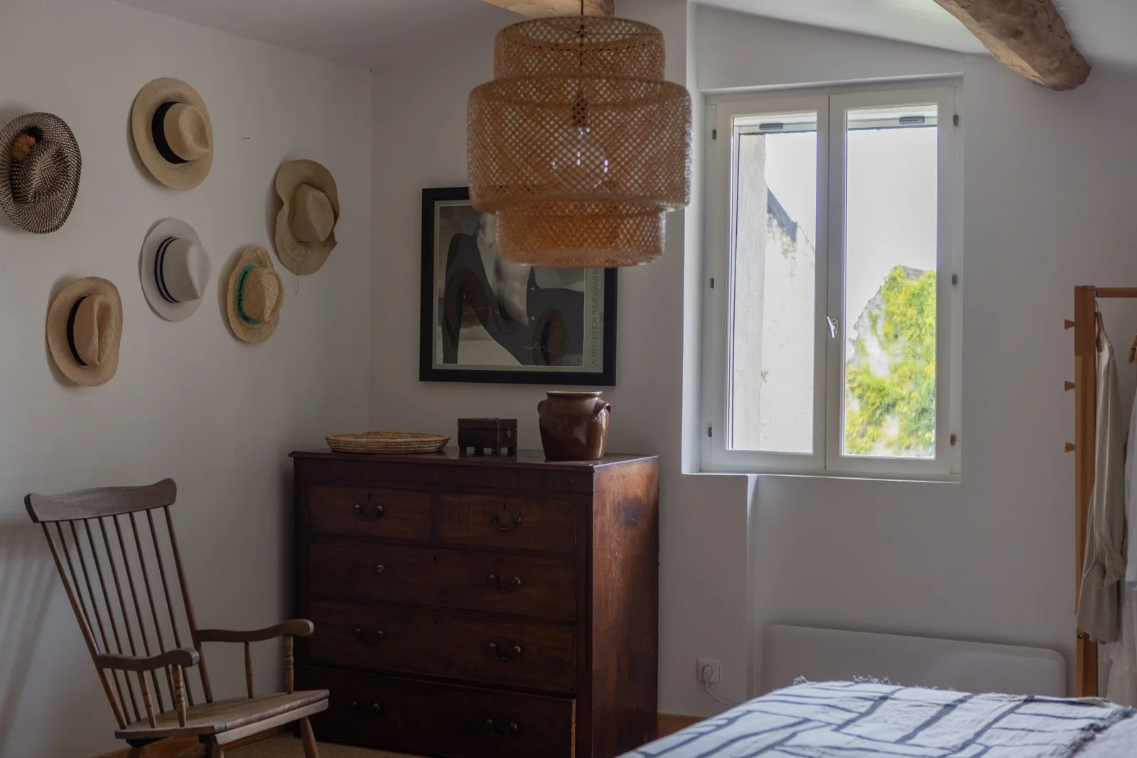 A bedroom with white walls, a wooden dresser, a wooden chair, a window showing green trees outside, wall-mounted hats, framed art on the wall, and a hanging woven lampshade.