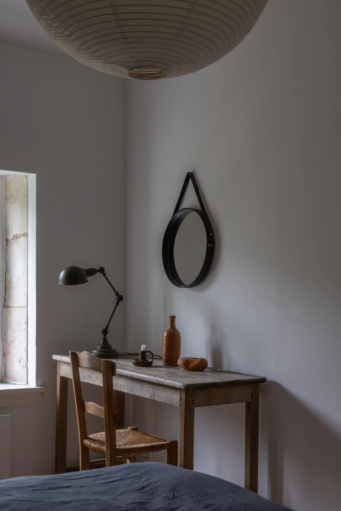 A small rustic wooden desk with a black lamp, a brown bottle, and decorative stones, situated near a window in a minimalist room with a mirror on the wall.