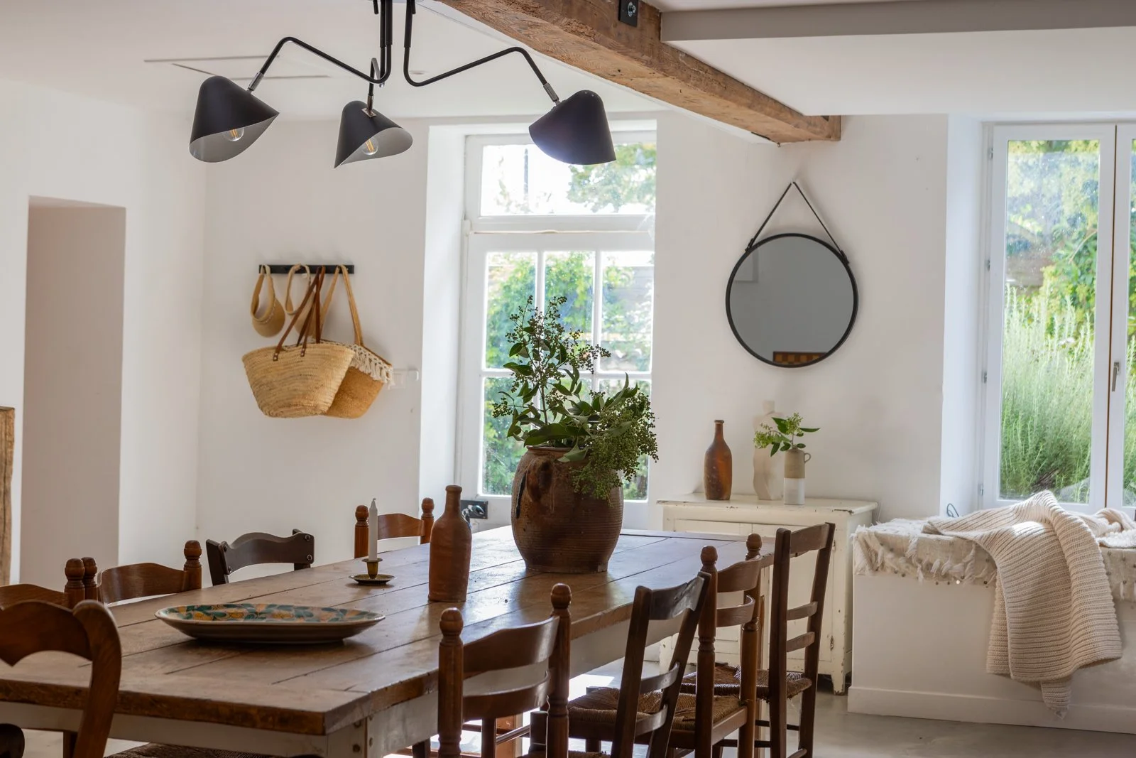 A rustic dining room with a wooden table, chairs, large vases, and natural decor, with windows bringing in natural light.