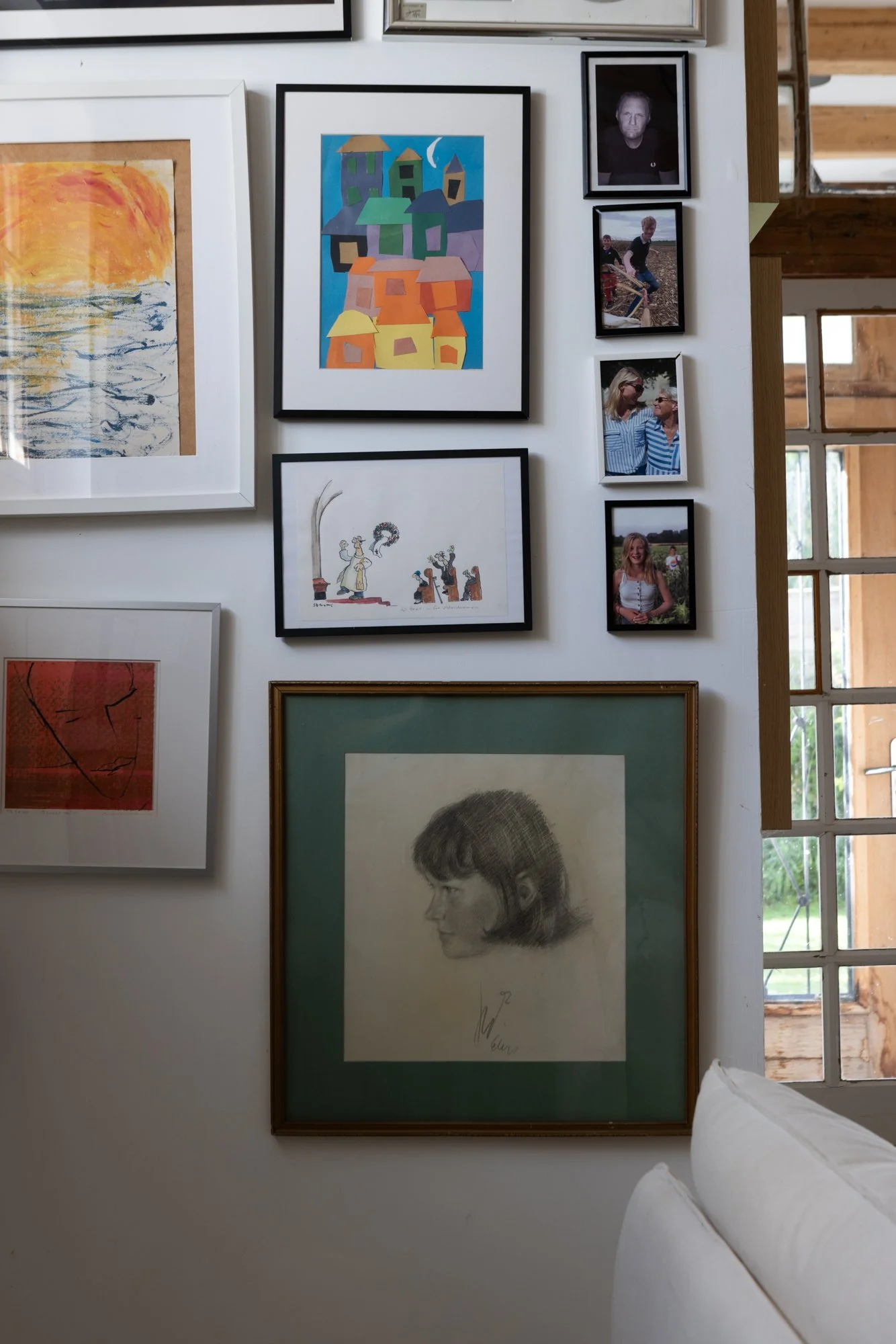 A collection of framed artwork and photographs on a white wall, including colorful paintings, a charcoal sketch of a woman with short hair, and smaller portraits of people, with a window on the right side allowing natural light.