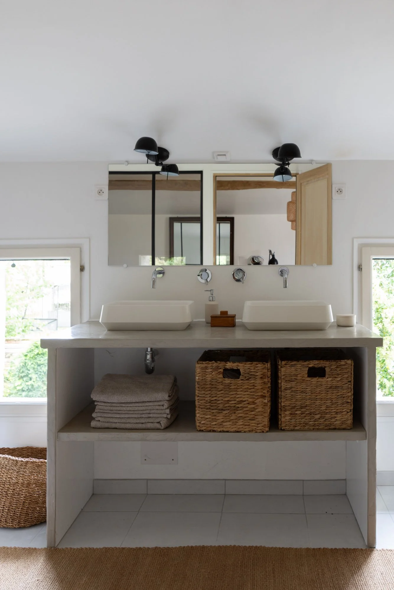 Bathroom with dual white vessel sinks, large mirror above, black wall sconces, towels in a shelf, woven baskets, windows with greenery outside, minimalist decor.