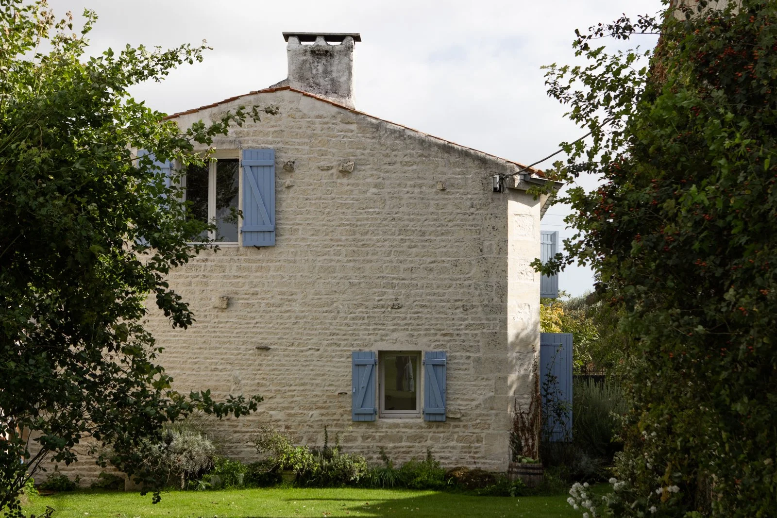 A stone house with blue shutters on windows, surrounded by greenery and trees.