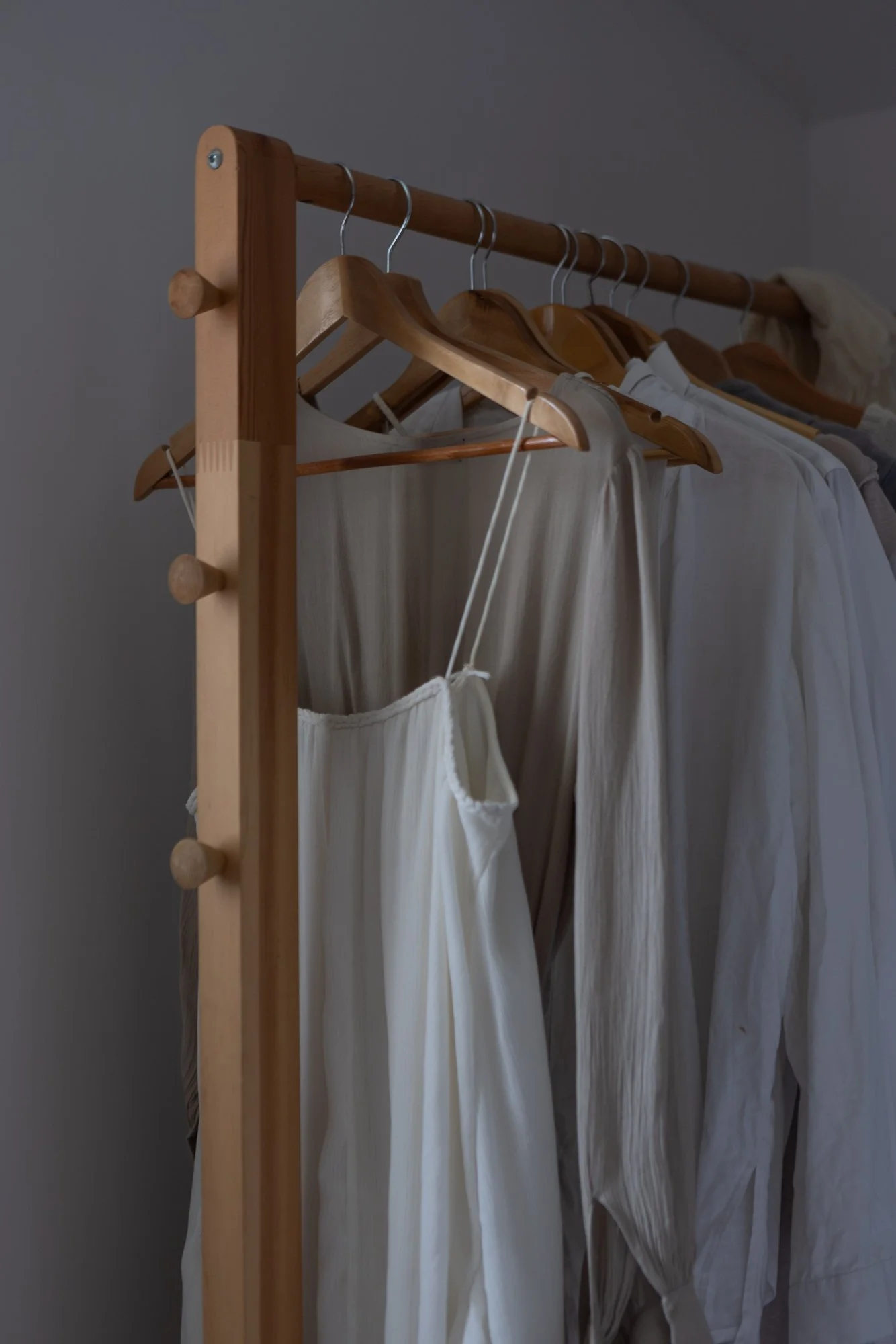 Wooden clothing rack with white and beige garments on wooden hangers, including a white sleeveless dress, in a dimly lit room.