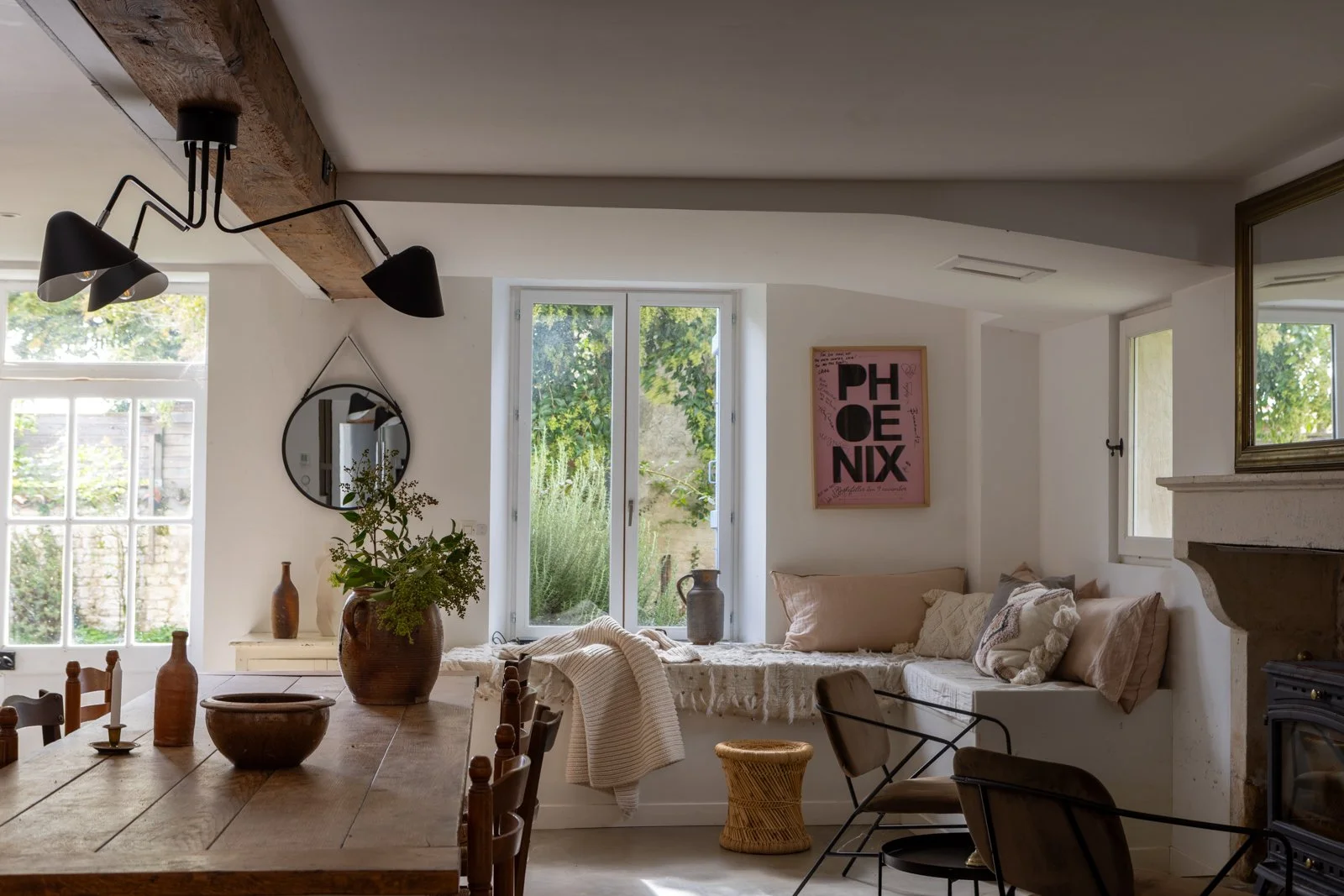 Cozy living room with a wooden dining table, decorative vases, a large potted plant, woven baskets, a wall mirror, artwork, window seat with cushions, and views of greenery outside.