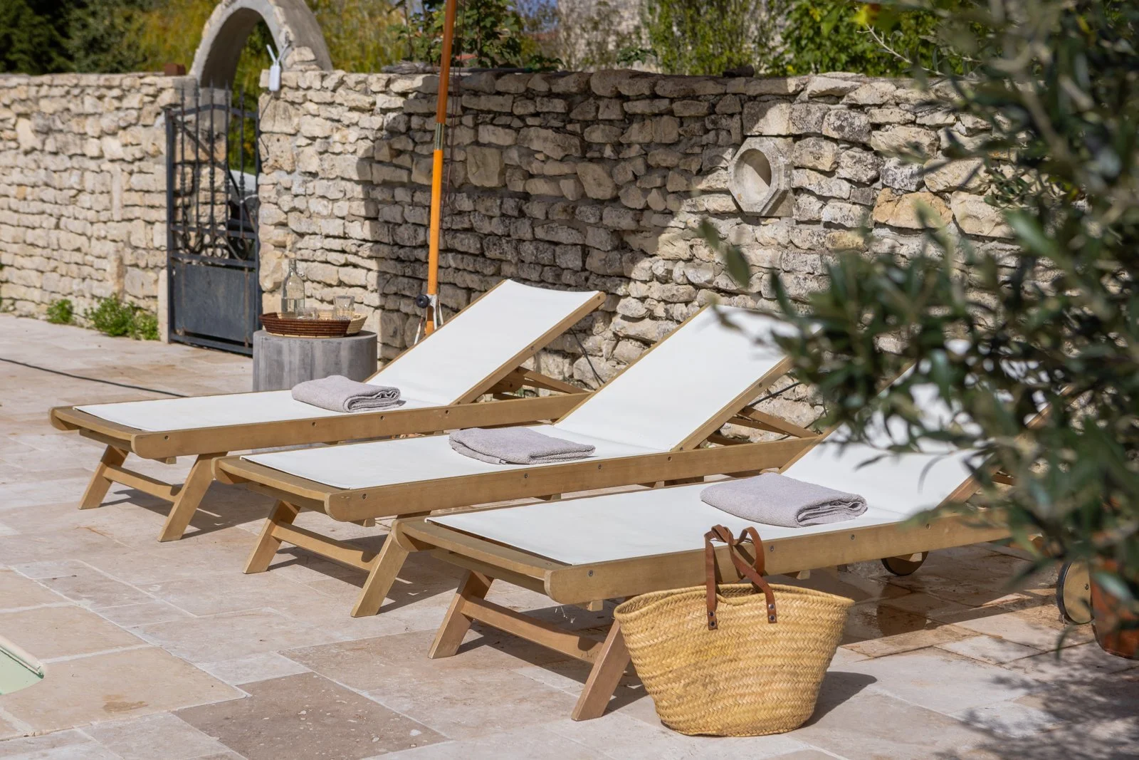 Two wooden lounge chairs with white cushions and gray towels on a stone patio, with a brick wall and greenery in the background.