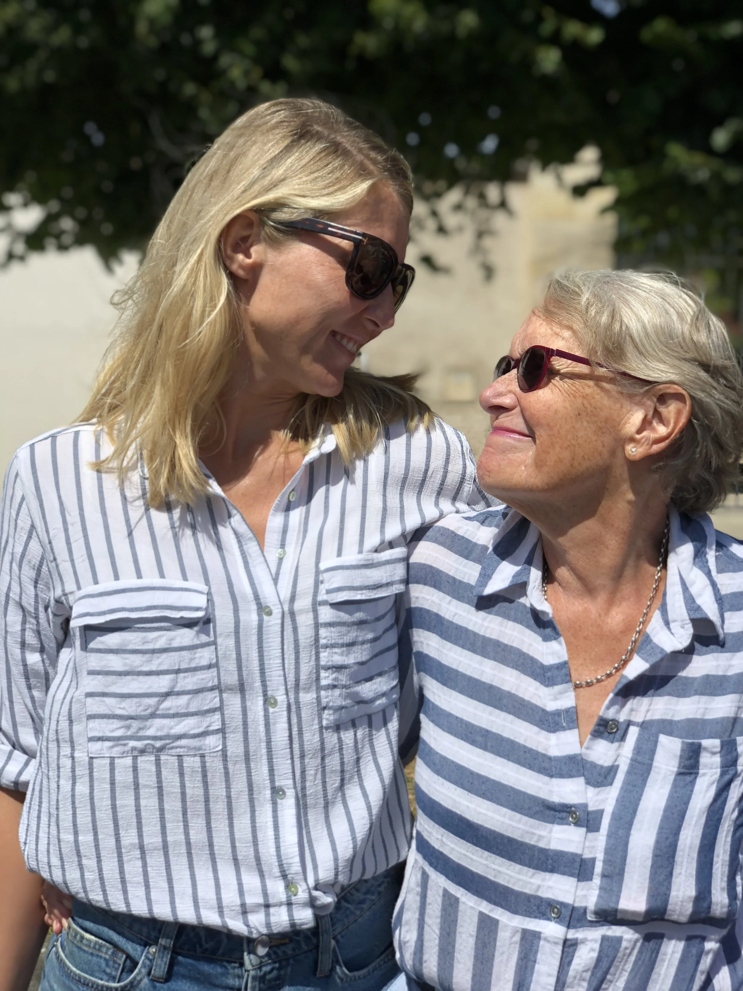 Two women, one younger and one older, smiling and looking at each other outdoors, both wearing sunglasses and striped shirts.