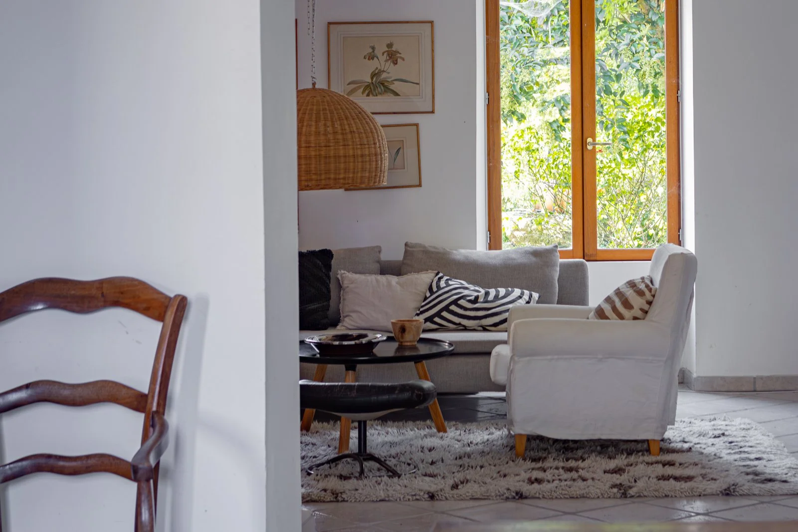 Cozy living room with gray sofa, white armchair, black and white striped pillow, wooden window with green foliage outside, decorative wall art, hanging wicker lamp, and a small black coffee table on a shaggy rug.