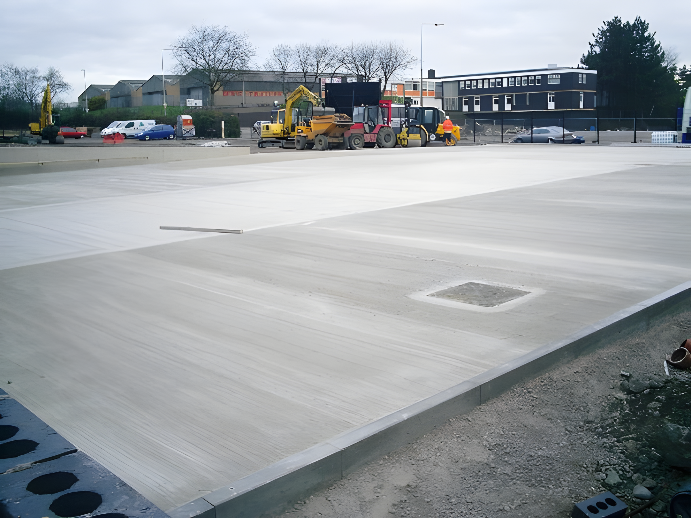Construction site with large flat surface covered in a white material, construction equipment, workers, and parked vehicles in the background.