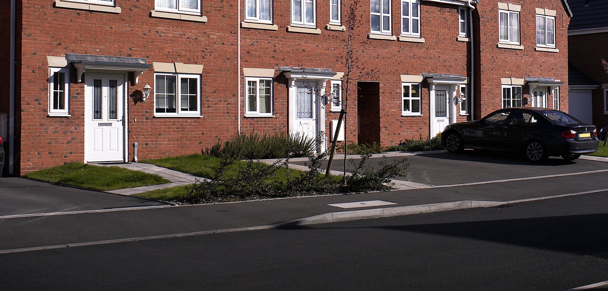 A row of brick residential townhouses with white doors and windows, a parking lot with a black car, and small garden patches in front.