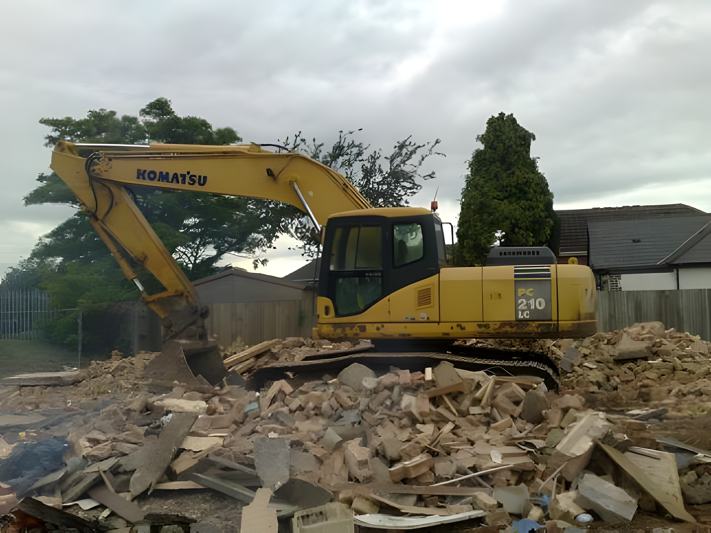 A yellow Komatsu excavator is working on a construction or demolition site, moving debris and rubble with a cloudy sky overhead.