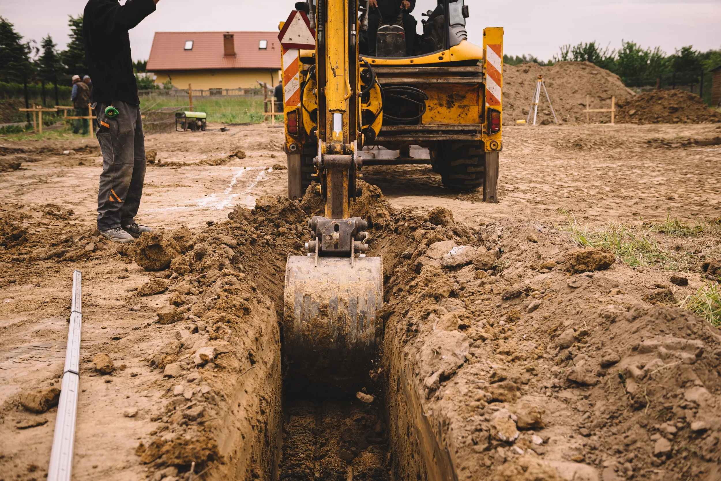 Construction site with a small yellow excavator digging a trench. A construction worker stands nearby with a measuring tape, and pointers and tools are visible in the background.