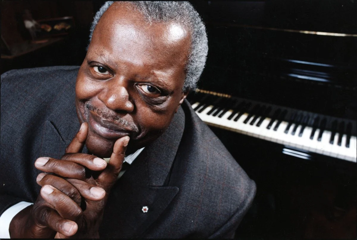 A black and white photo of Oscar Peterson playing a piano, wearing a light-colored suit, with an audience in the background. The photo has a signature in blue ink signing the image.