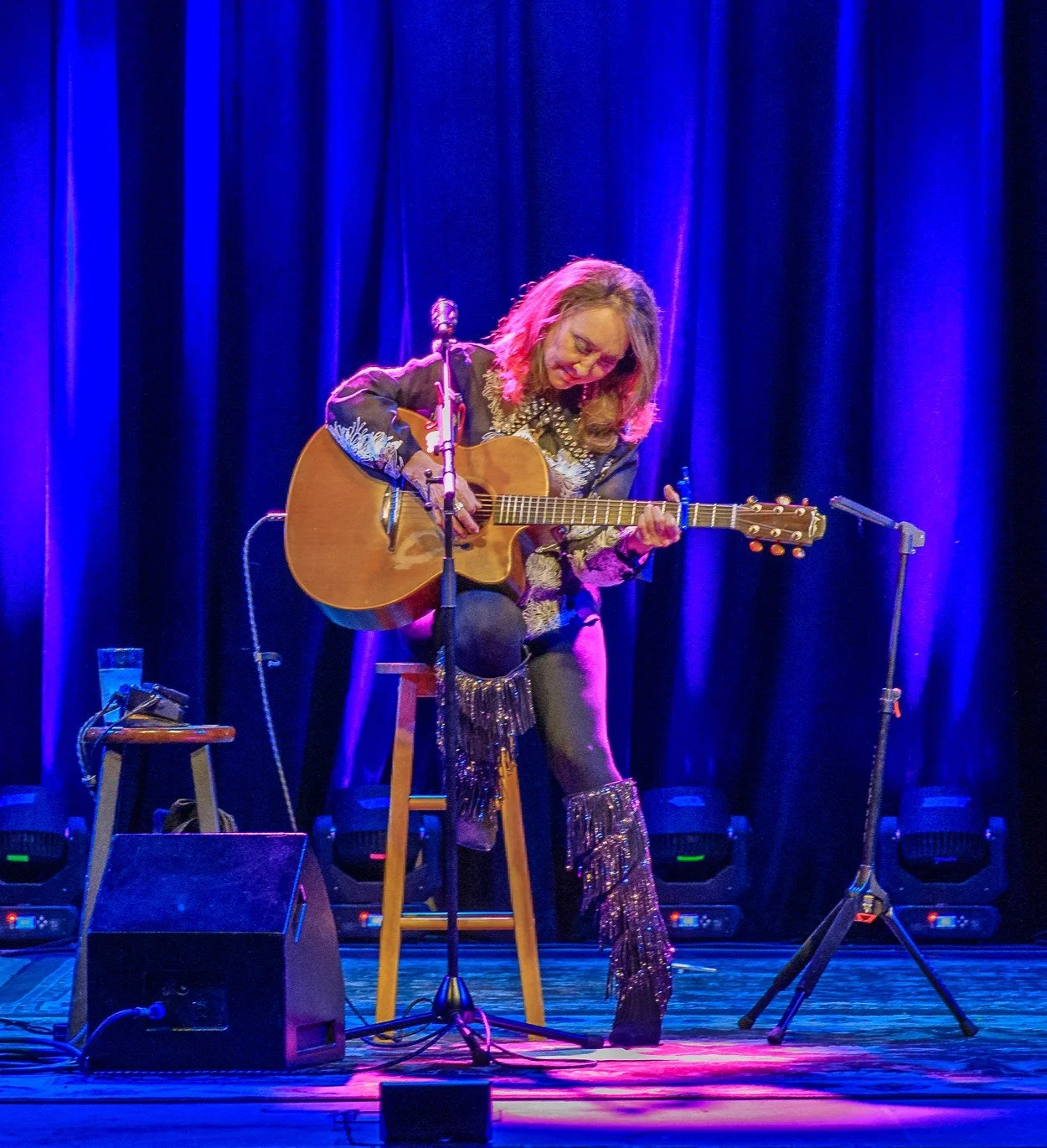 Digital artist, Concert lighting director, writer Woody Bavota, Pam Tillis playing acoustic guitar on stage with dark blue curtains, stage lighting, and audience in foreground.