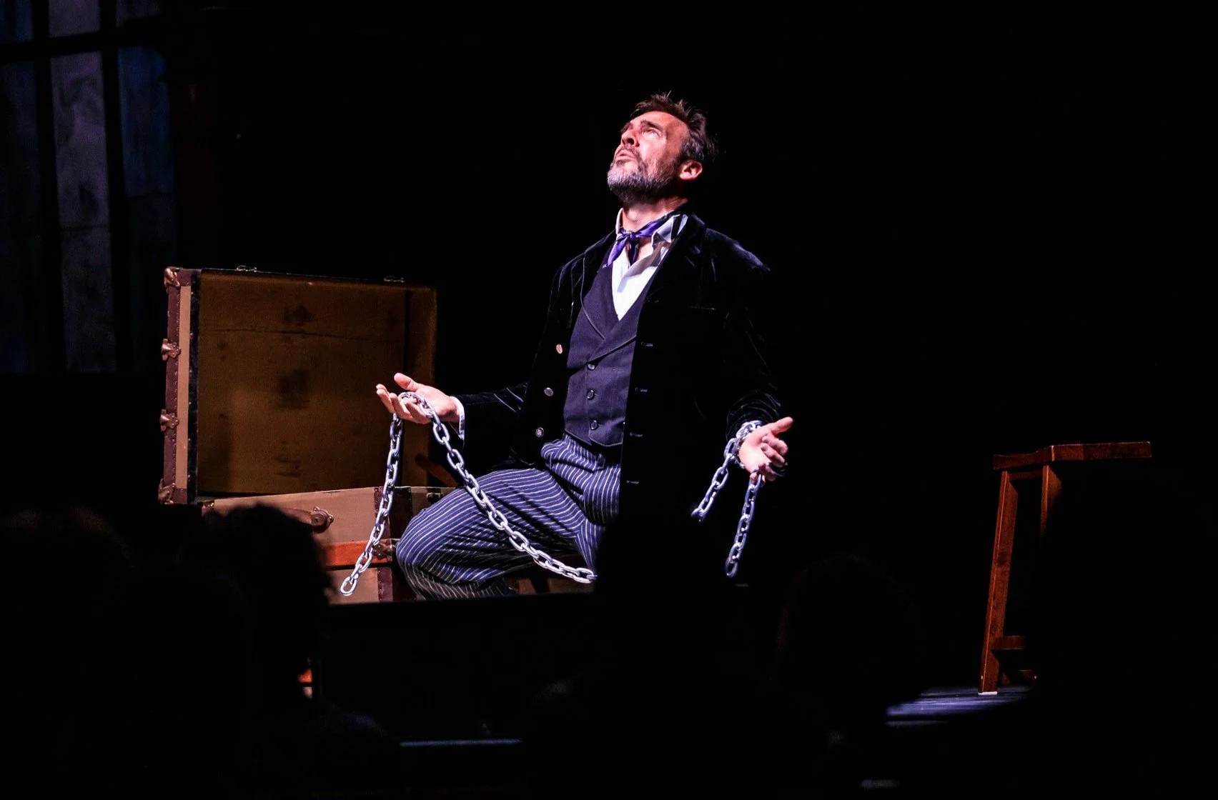 Digital artist, Concert lighting director, writer Woody Bavota, Man with light-colored hair, wearing a black jacket, striped pants, and shiny shoes, jumping with arms raised and mouth open on a dark stage.
