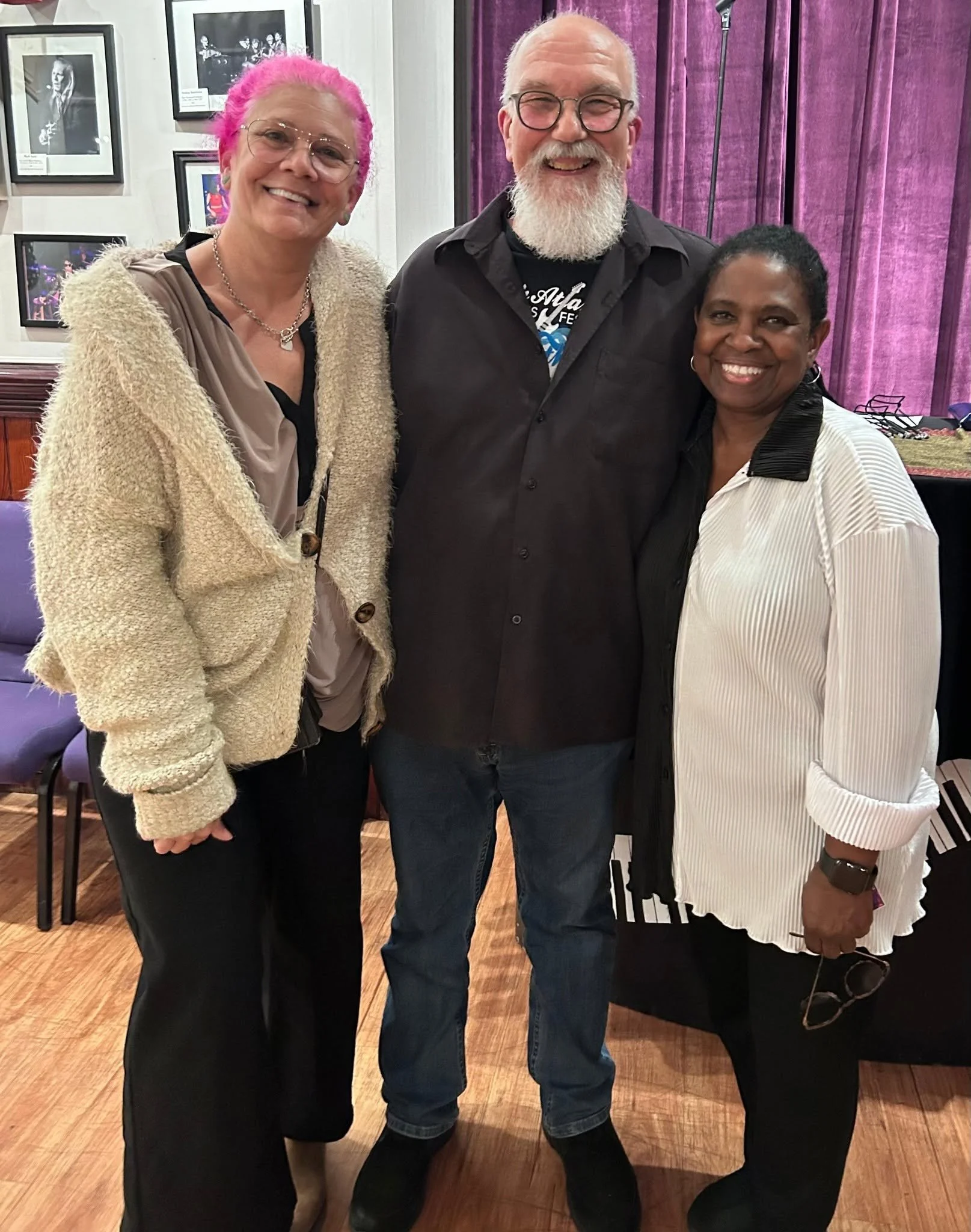Three smiling people standing together indoors, posing for the photo, with a dark purple curtain background.
