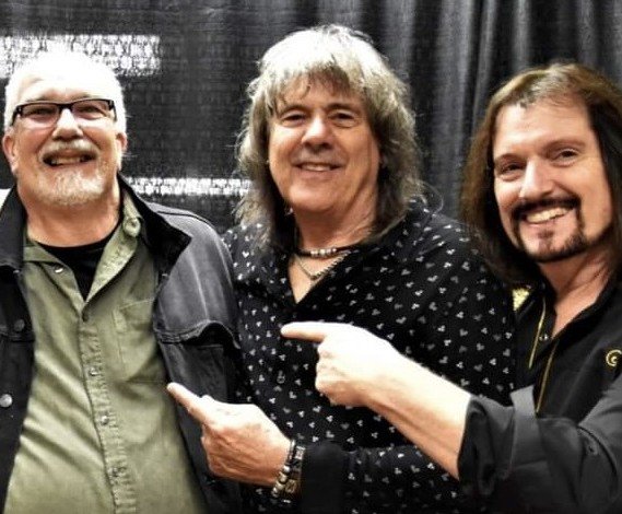 The band New England and Woody standing together, smiling, with a black curtain background. Woody Bavota digital artist, concert lighting, writer.