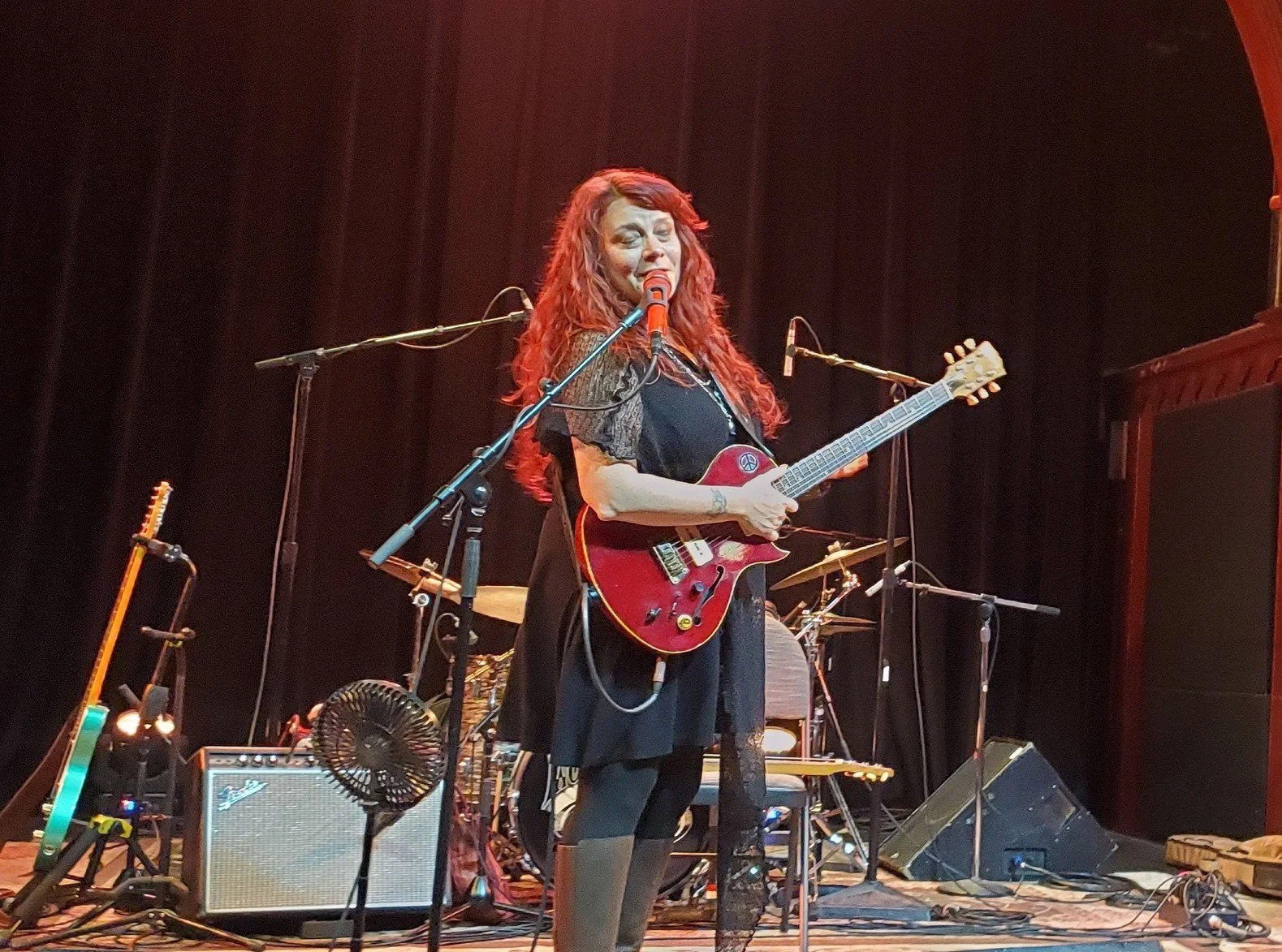 Digital artist, Concert lighting director, writer Woody Bavota, A woman with long red hair passionately singing into a microphone while playing an electric guitar, under red stage lighting.