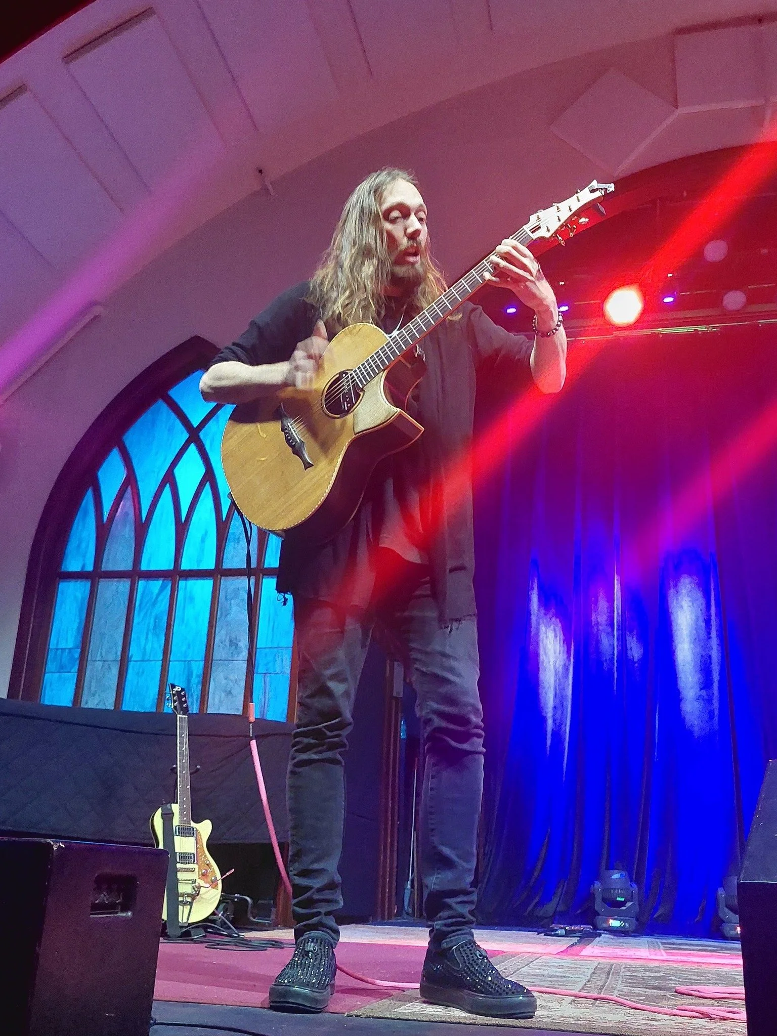 Digital artist, Concert lighting director, writer Woody Bavota, A musician with long, curly hair playing an acoustic guitar on stage illuminated by colorful stage lights, wearing a black shirt and dark pants.