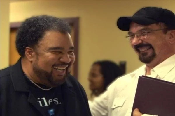 Two men smiling and talking to each other, with a woman in the background.