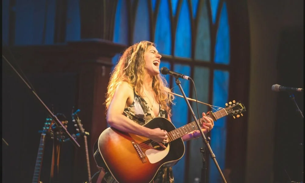 Digital artist, Concert lighting director, writer Woody Bavota, Sarah Martin performing on stage with a guitar, surrounded by purple and blue stage lighting.