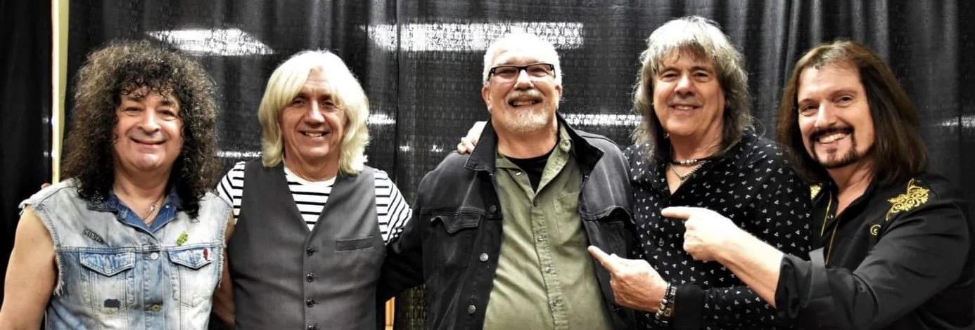 The band New England and Woody standing together, smiling, with a black curtain background. Woody Bavota digital artist, concert lighting, writer.