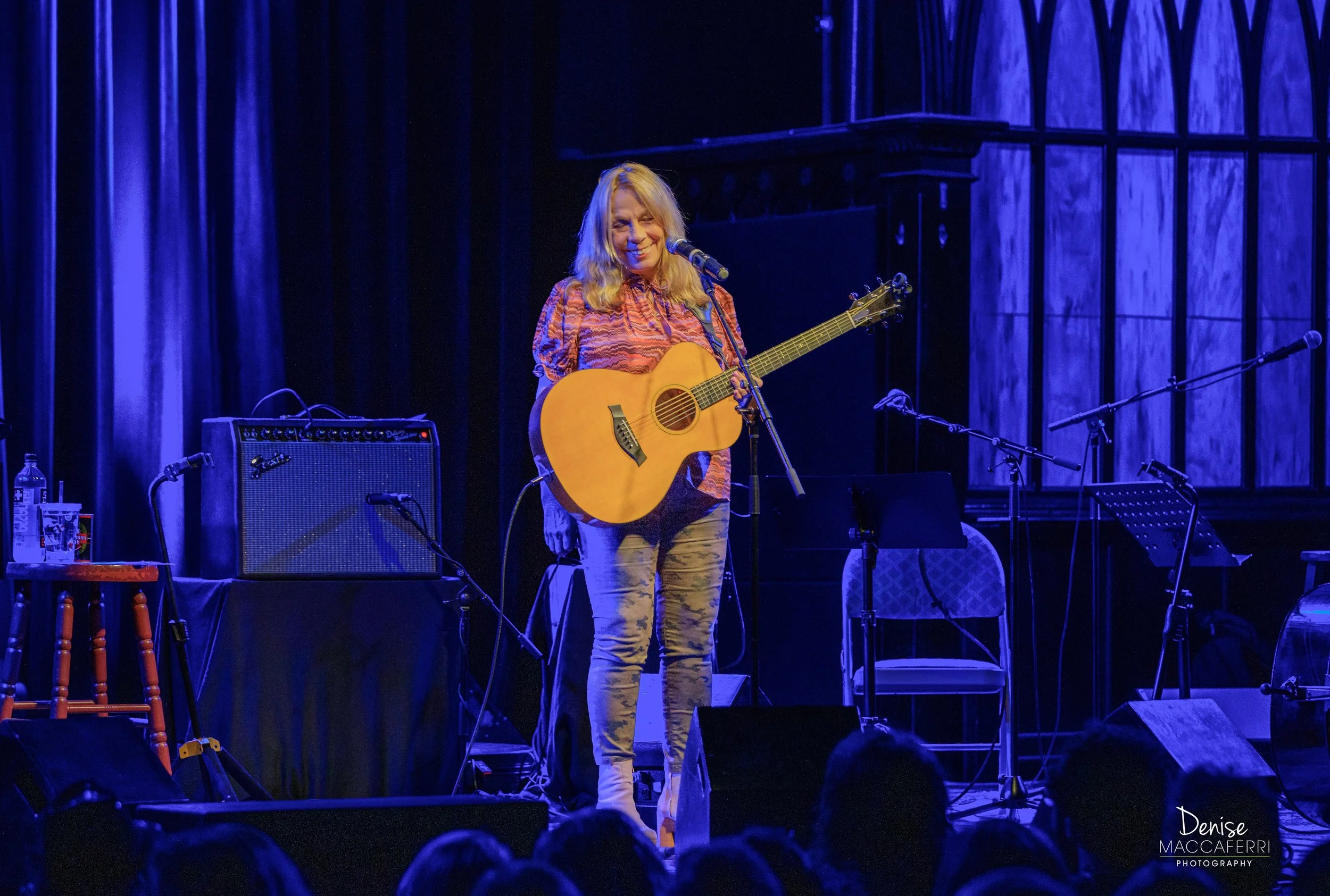 Digital artist, Concert lighting director, writer Woody Bavota, Rickie Lee Jones on stage playing an acoustic guitar and singing into a microphone, with blue stage lighting and musical equipment around her.