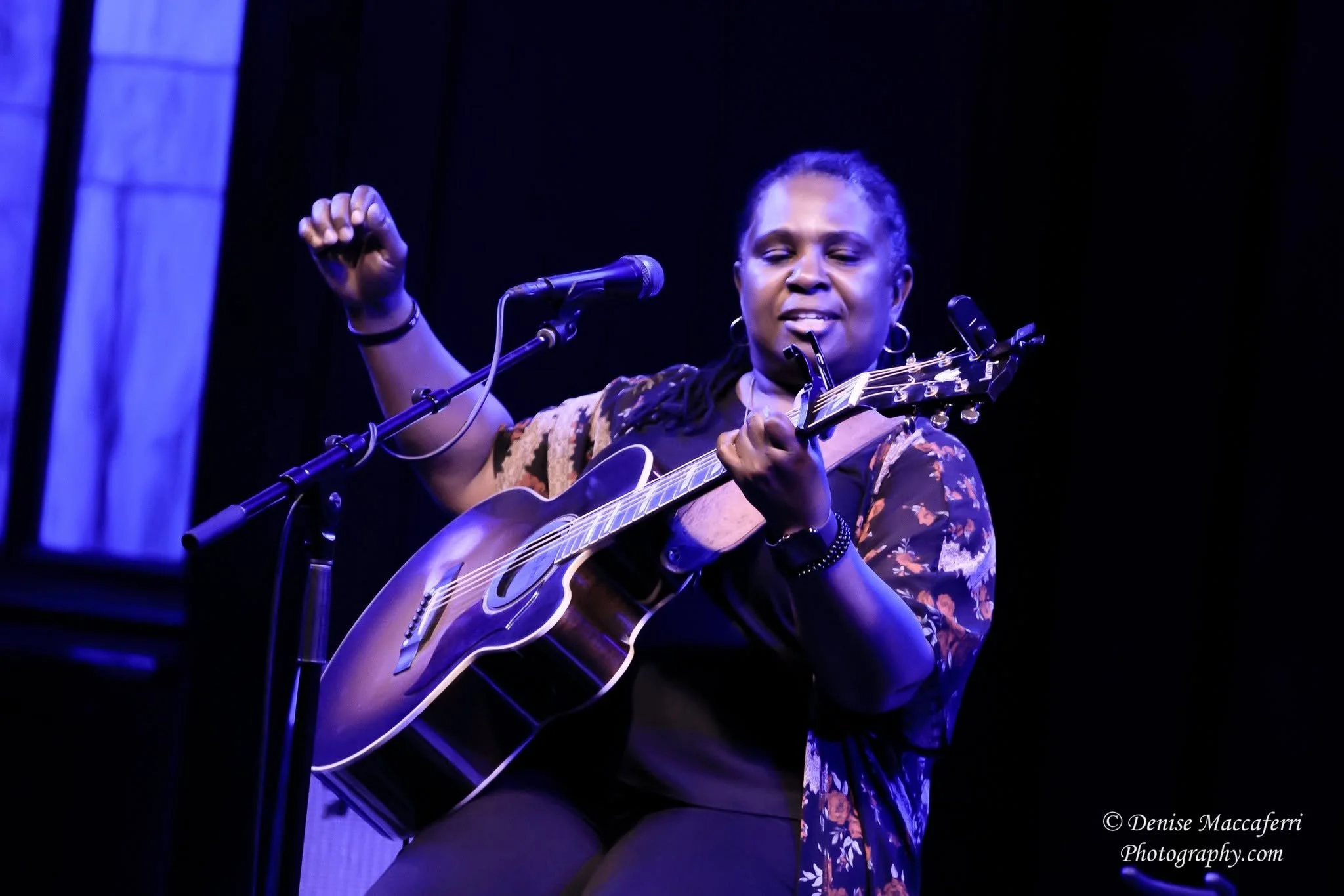 Digital artist, Concert lighting director, writer Woody Bavota, A woman with braids and jewelry plays an acoustic guitar and sings into a microphone on a dark stage, eyes closed and emotional.