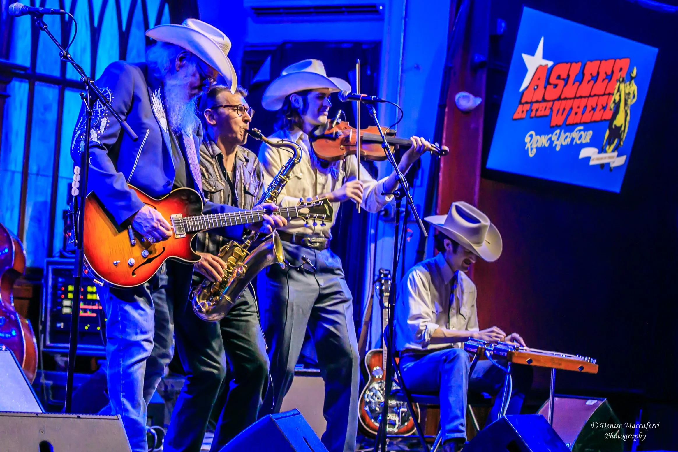 A country band performing on stage with five band members, wearing cowboy hats, playing guitar, saxophone, drums, double bass, and keyboard. Audience seated in front of stage, some taking photos.