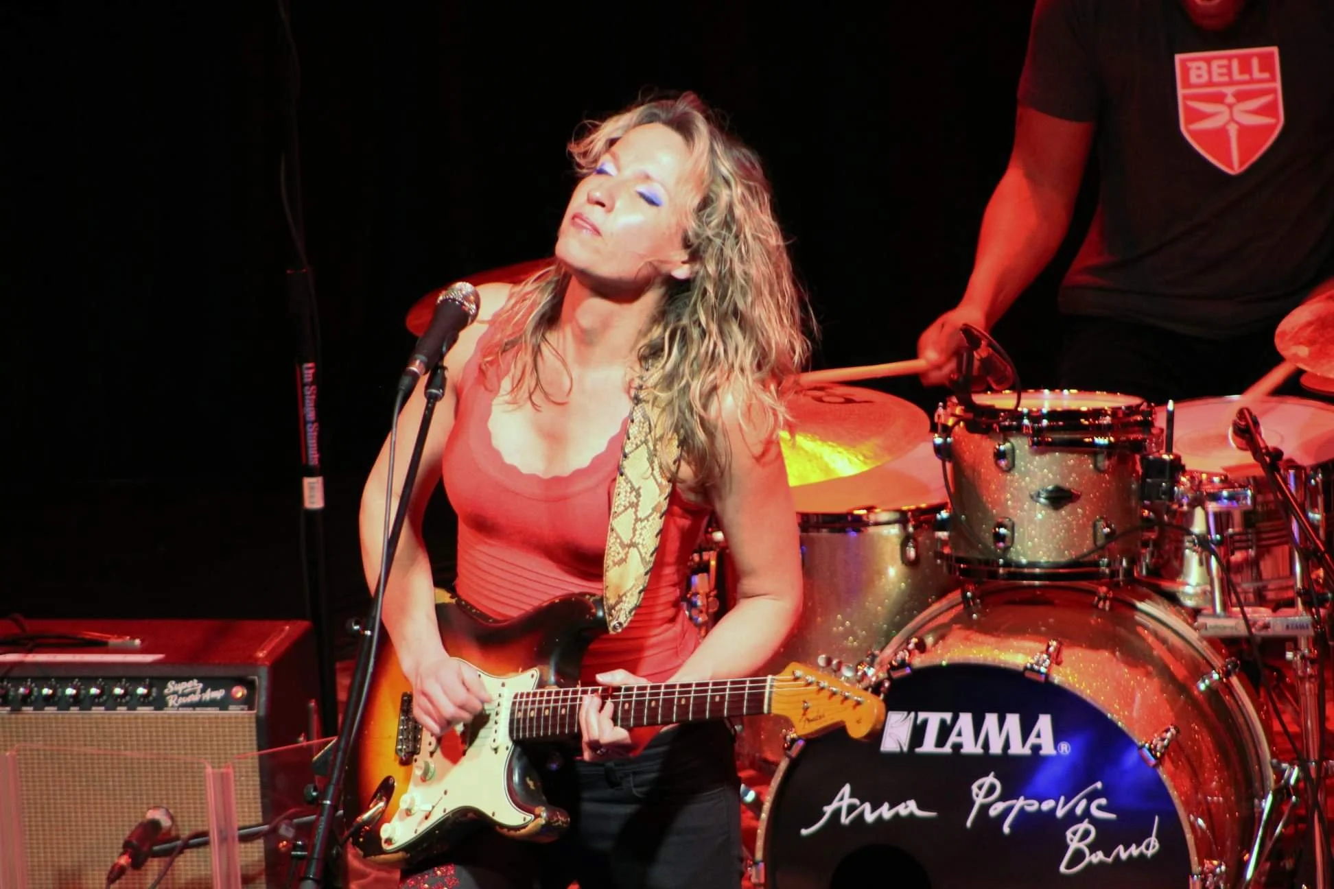 Female musician playing electric guitar on stage with drums in the background, featuring a Tama logo and signature of Anna Popovic Band.