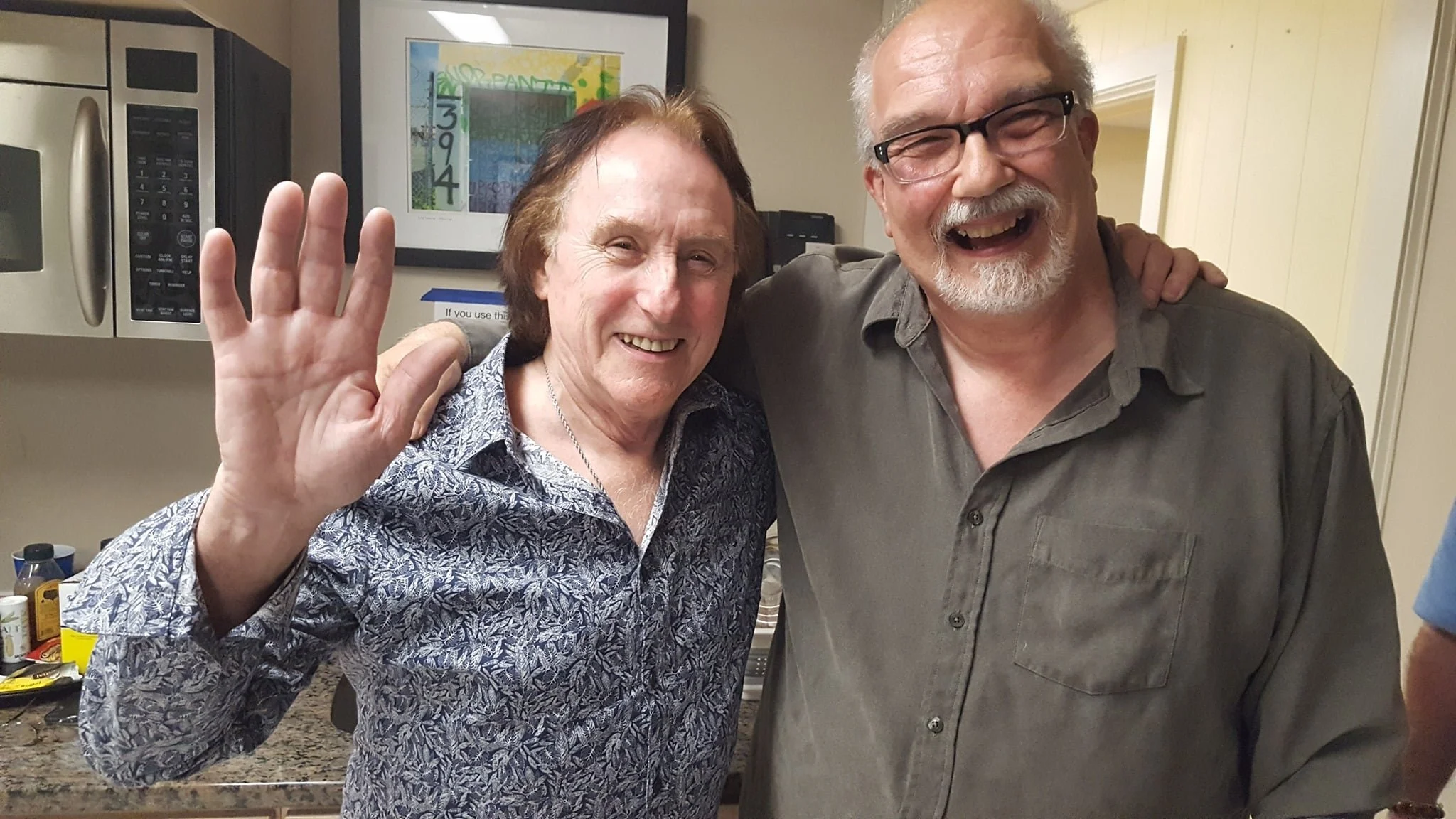 Two older men smiling and laughing, one with his arm around the other, waving at the camera, in a kitchen setting.
