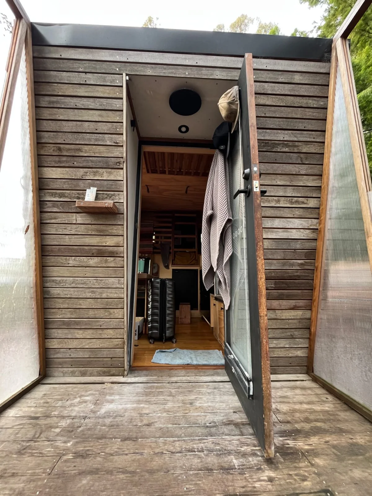View inside a tiny wooden house with an open door, showing a small living space with luggage, hanging towels, and wooden furniture.