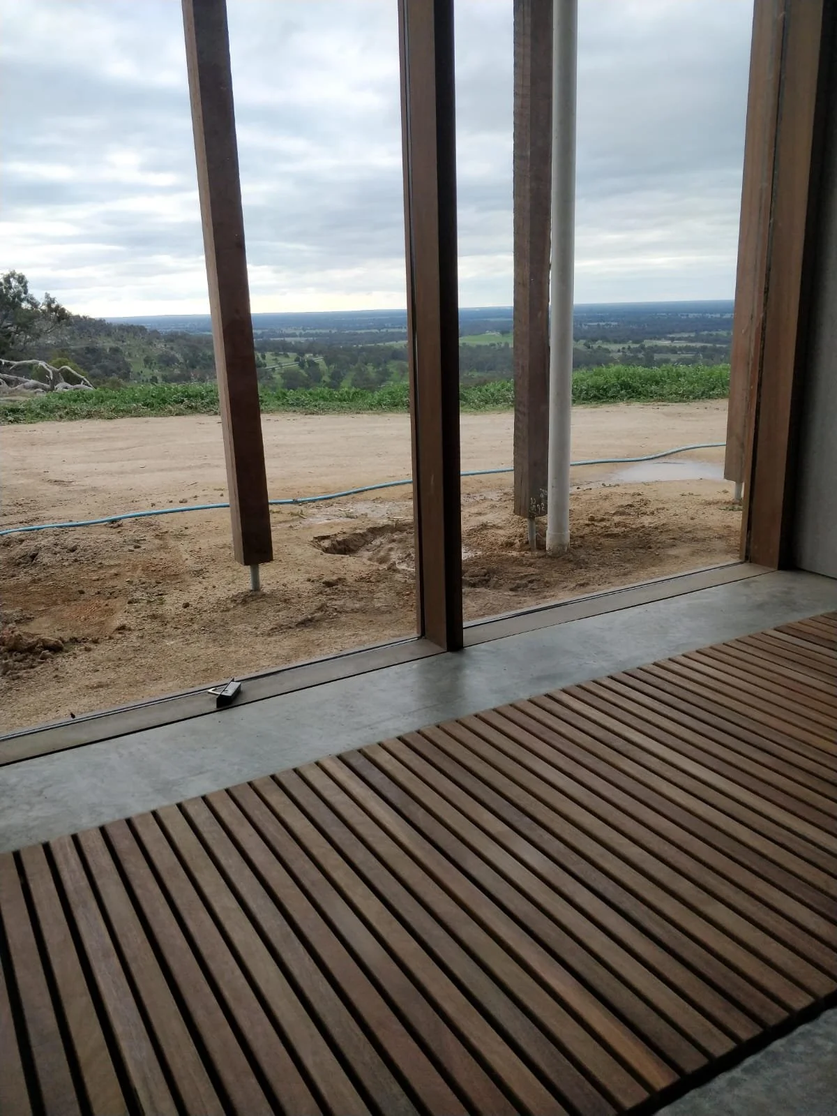 View through a glass sliding door showing a dirt pathway and green fields with rolling hills and cloudy sky in the distance.