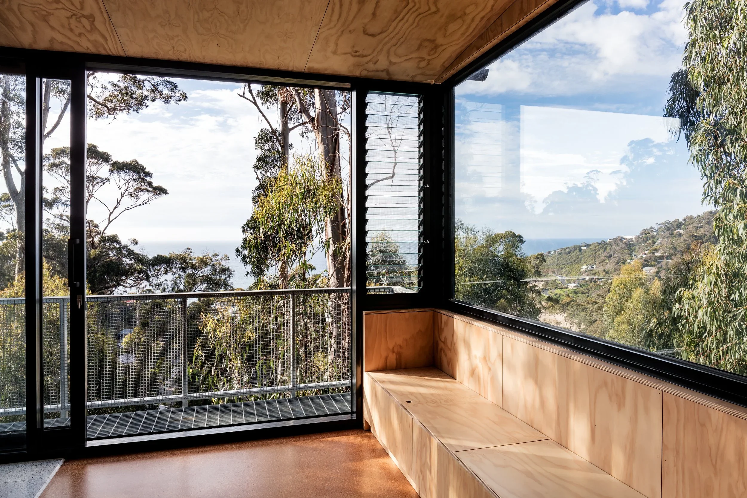 Interior view of a room with large glass windows and a glass sliding door, overlooking a lush green landscape with trees and a distant view of the sea or sky.