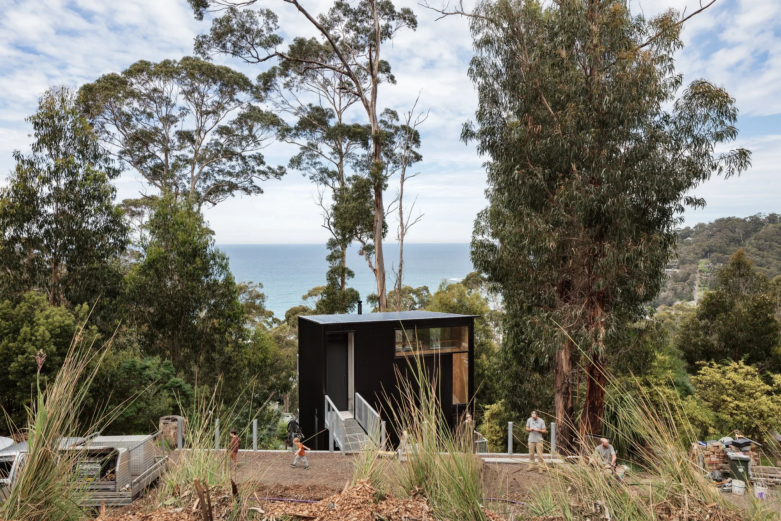 Modern black house situated on a hillside among tall trees, overlooking a view of the sea in the distance. Construction work is visible with workers on site.