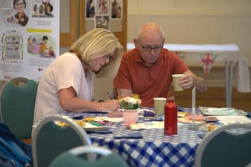 An elderly man and woman sitting at a table with a blue and white checkered tablecloth, engaged in an activity, with cups and decorations on the table.