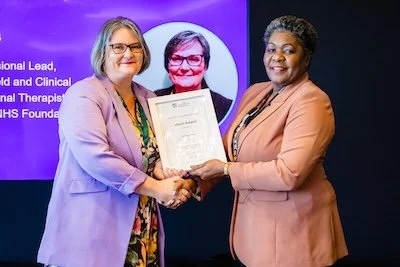 Two women shaking hands while holding a certificate, with a purple background and a portrait of a woman in a circle on the screen behind them.