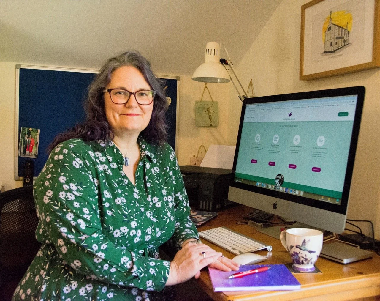 A woman with glasses and long dark hair, wearing a green floral dress, seated at a desk with her hands on a purple notebook, smiling at the camera. On her desk, there is a white keyboard, a mug with a bird design, a red pen, and a computer monitor displaying a website. The background shows a blue notice board, a framed picture and some wall hangings.
