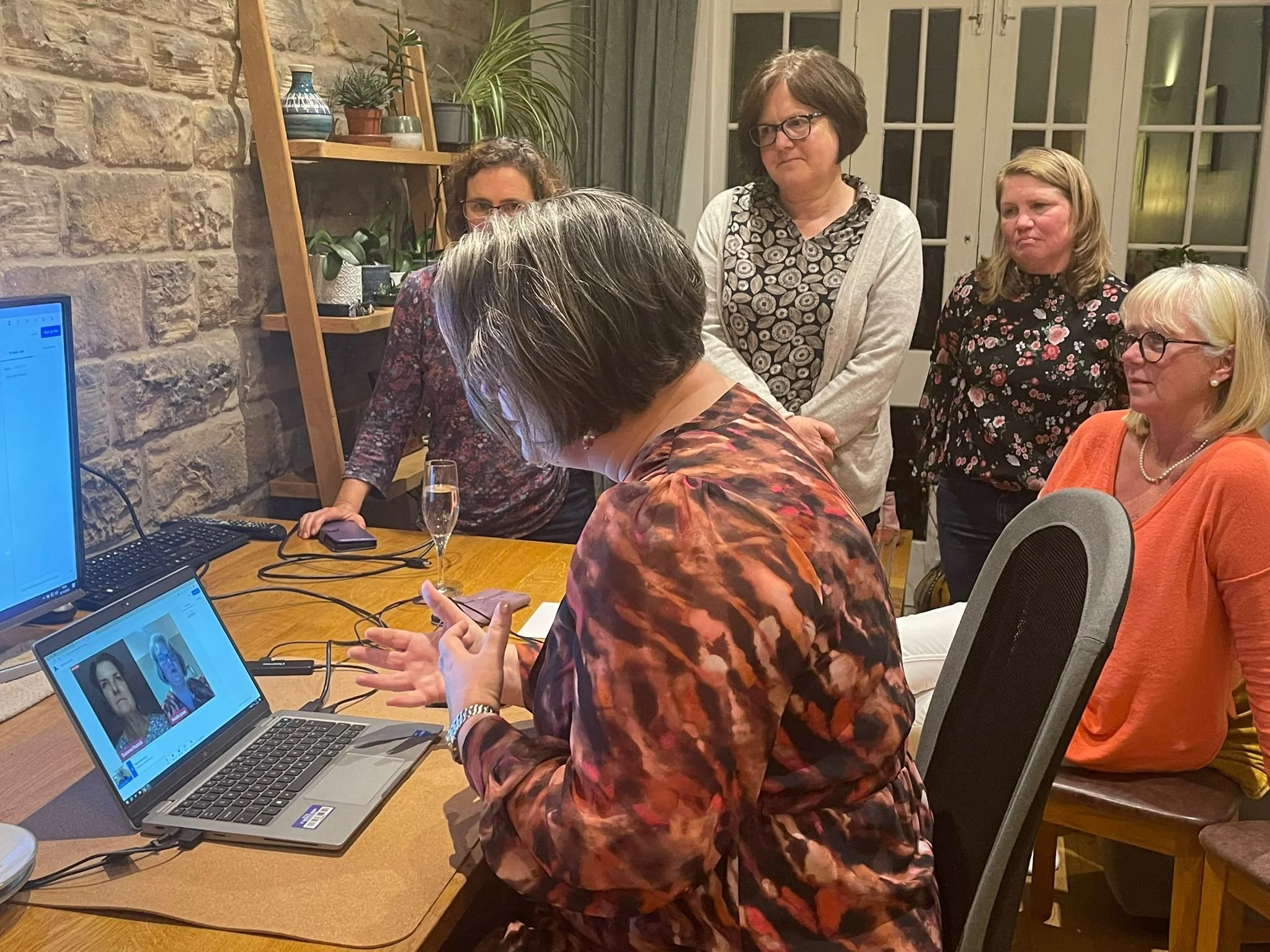 A group of six women gathered around a wooden table in a room, with one woman showing something on her phone. There are two laptops, one with a video call displayed, and a glass of champagne on the table. The room has a stone wall, plants, and a window with glass doors.