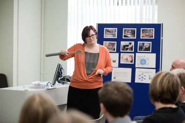 A woman with short dark hair and glasses, holding a pointer, standing in front of a blue presentation board with photos and charts, speaking to an audience in a room with white curtains.