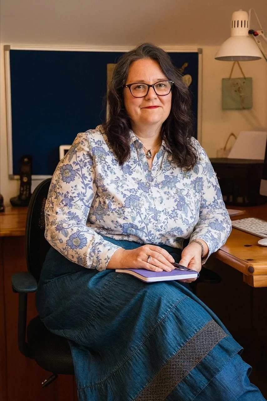 A woman with dark, wavy hair, glasses, wearing a floral blouse and a denim skirt, sitting on an office chair in an office setting, holding a notebook and pen.