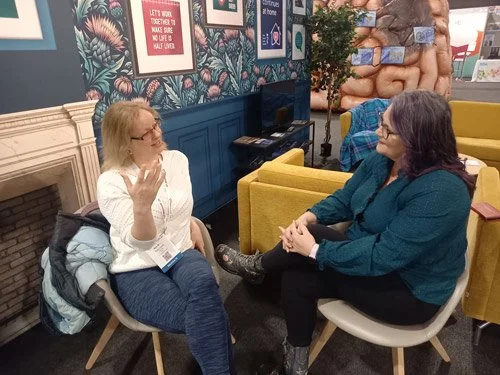 Two women sitting in chairs and talking in a cozy, decorated room with colorful walls, framed pictures, and a fireplace.