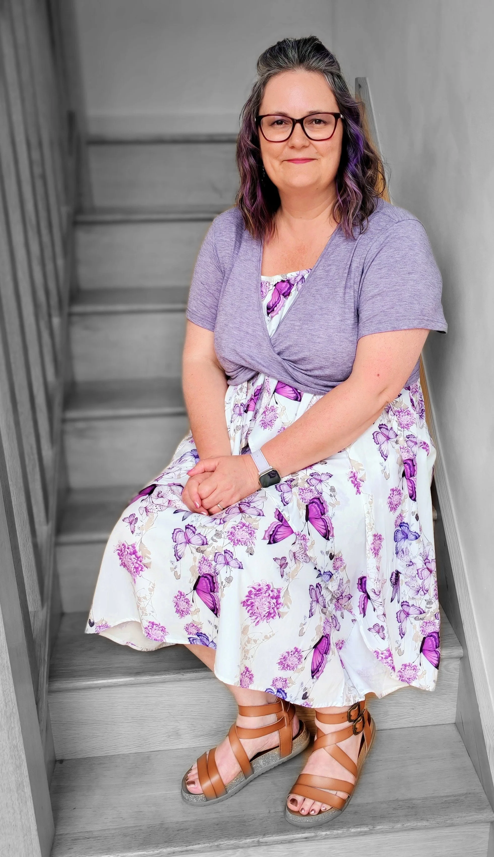 A woman sitting on a staircase, wearing glasses, a purple cardigan, a floral dress with purple butterflies, and tan sandal wedges.