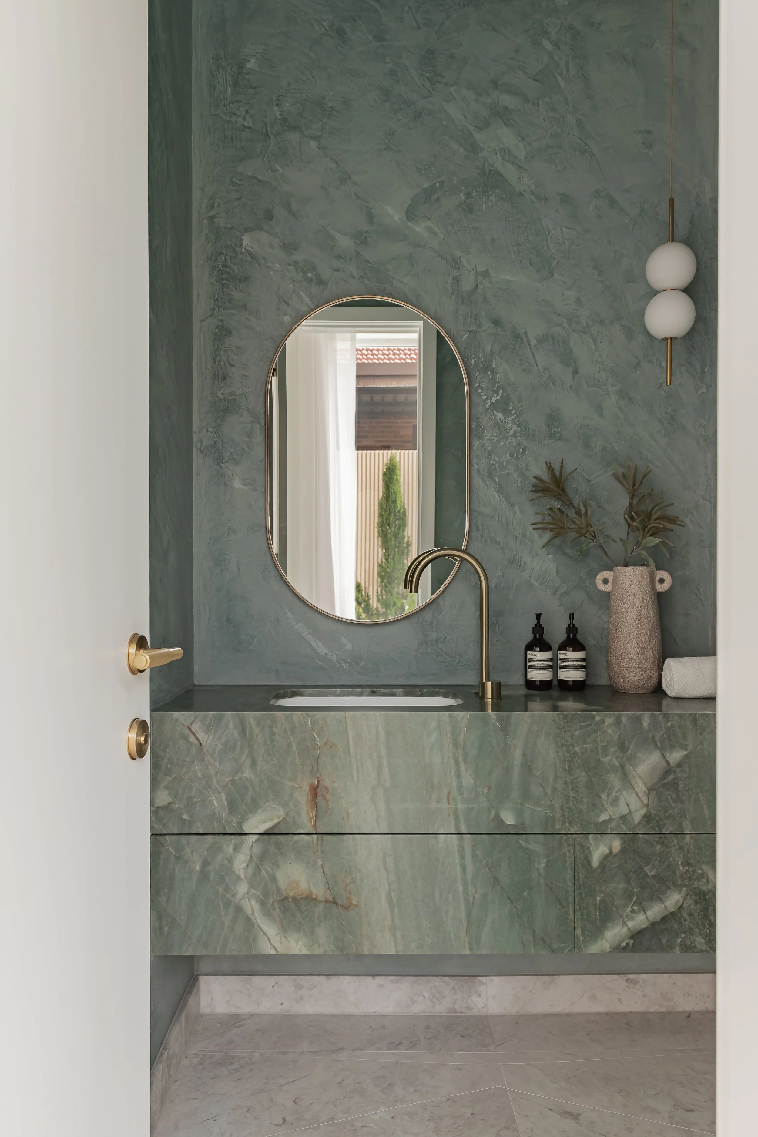 A modern bathroom featuring a green marble vanity with a built-in sink and a gold faucet. Above the vanity is an oval mirror, and on the right side, a textured grey vase with green leaves. There are two black bottles and a white towel on the surface, and a white wall-mounted light fixture with two spherical bulbs on the right side.
