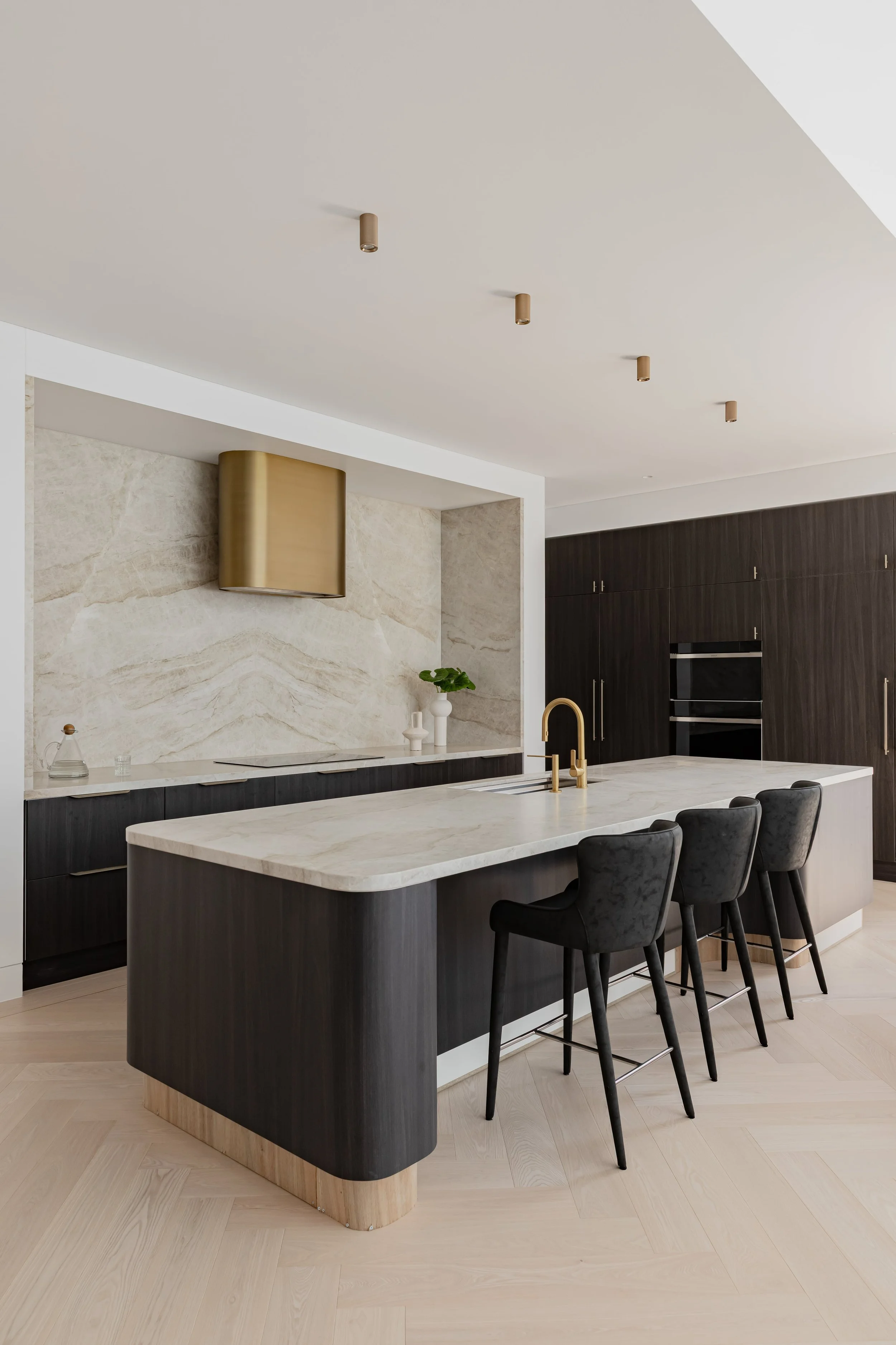 Modern kitchen with black cabinets, marble island, gold fixtures, and light hardwood floors.