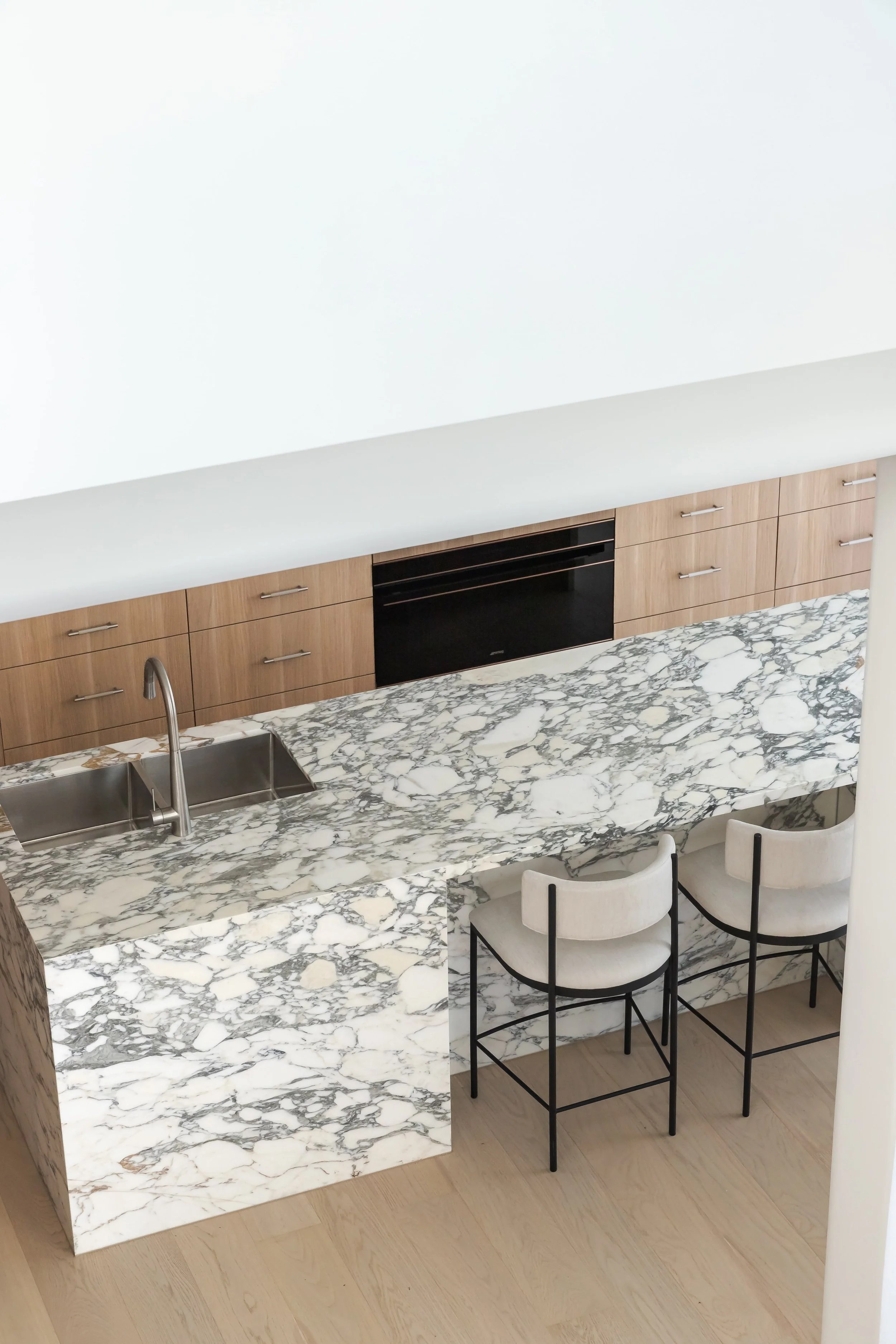 A modern kitchen with a marble countertop and backsplash, two bar stools, wooden cabinets, and a built-in oven.