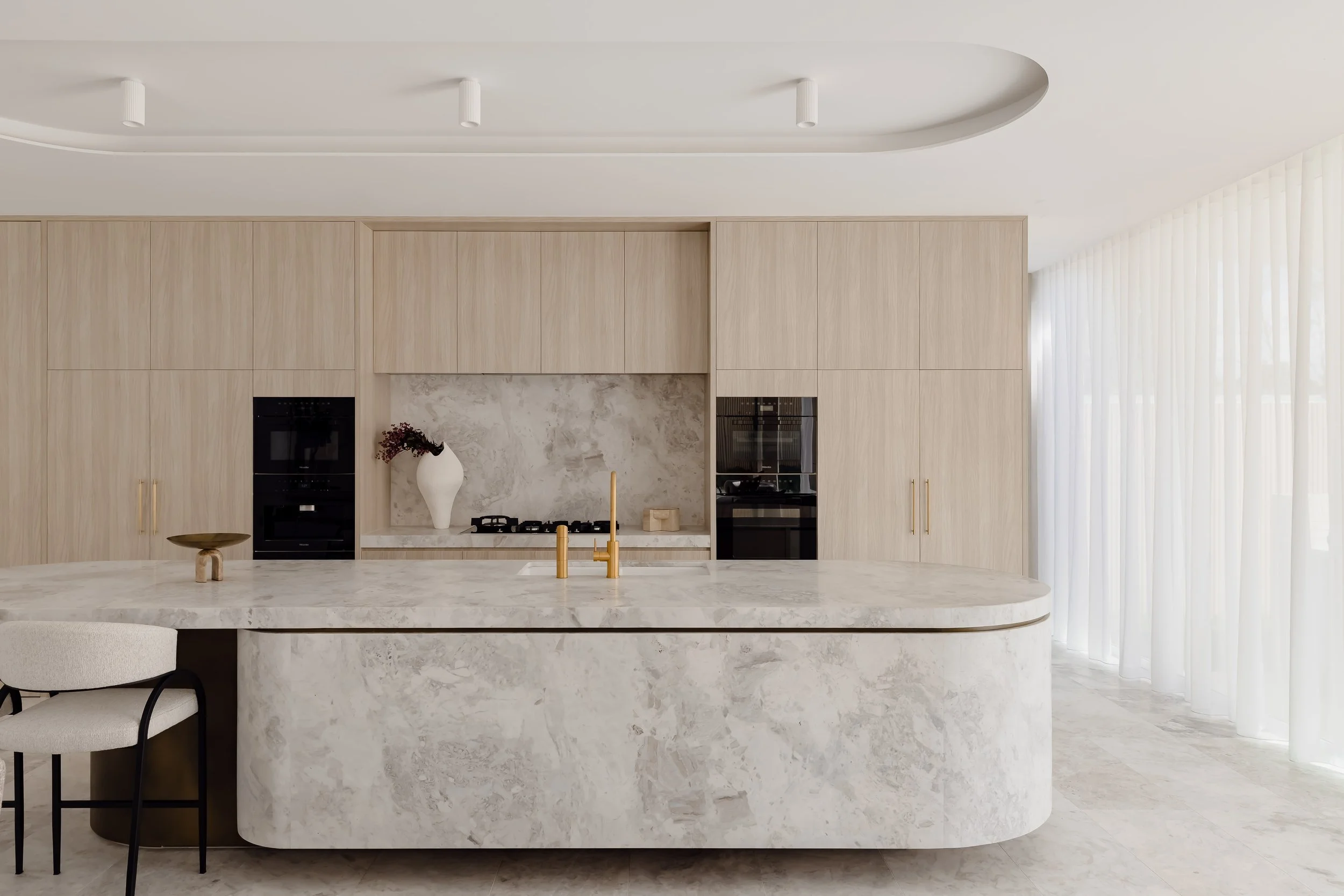 Modern kitchen with light wood cabinets, a large marble island with a built-in sink and gold faucet, and built-in black ovens. White curtains cover large windows on the right.