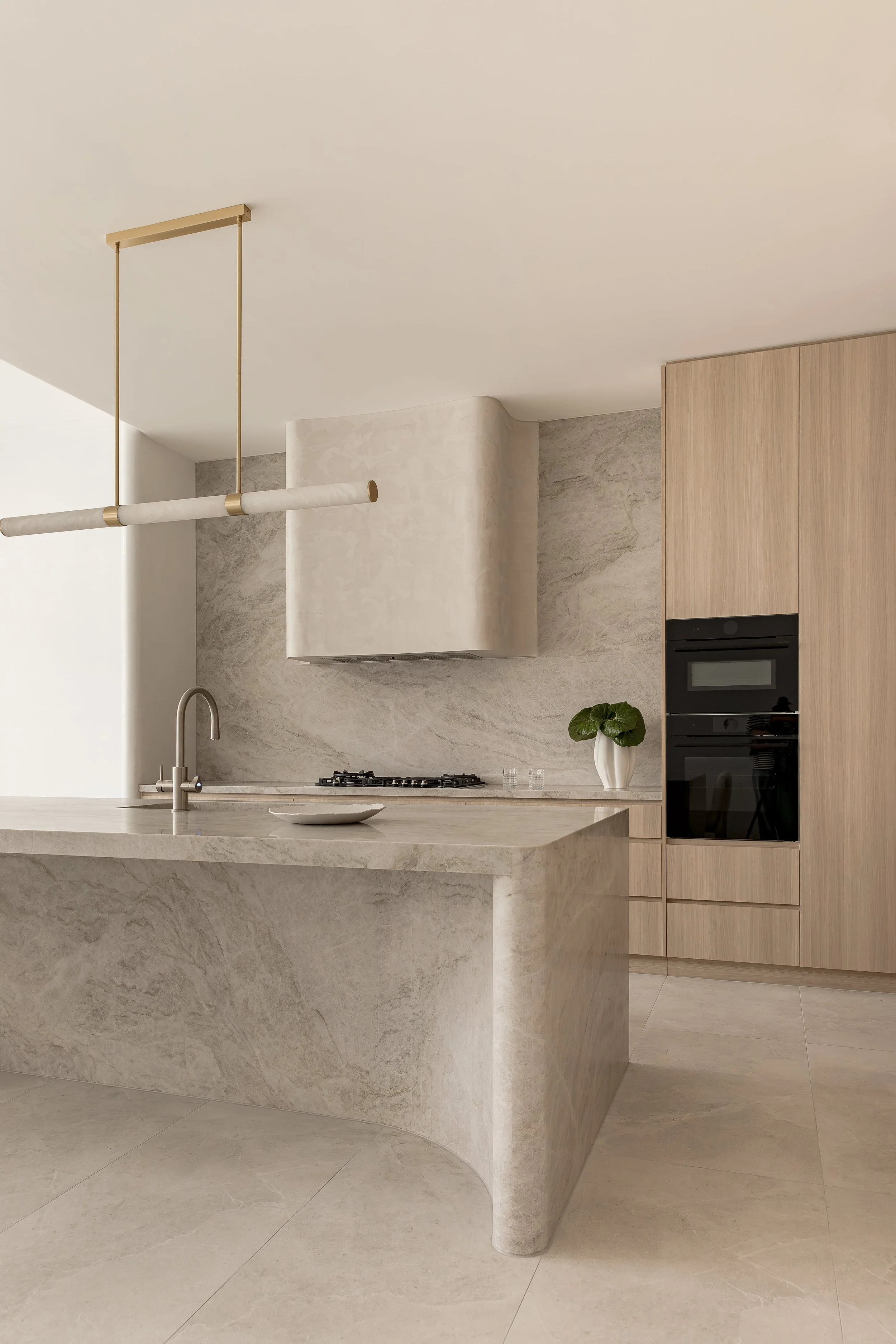 Modern kitchen with light wood cabinetry, a marble island, and a marble backsplash. A plant in a white vase is on the counter, and a sleek black microwave and oven are built into the cabinetry. A contemporary gold and marble pendant light hangs from the ceiling.