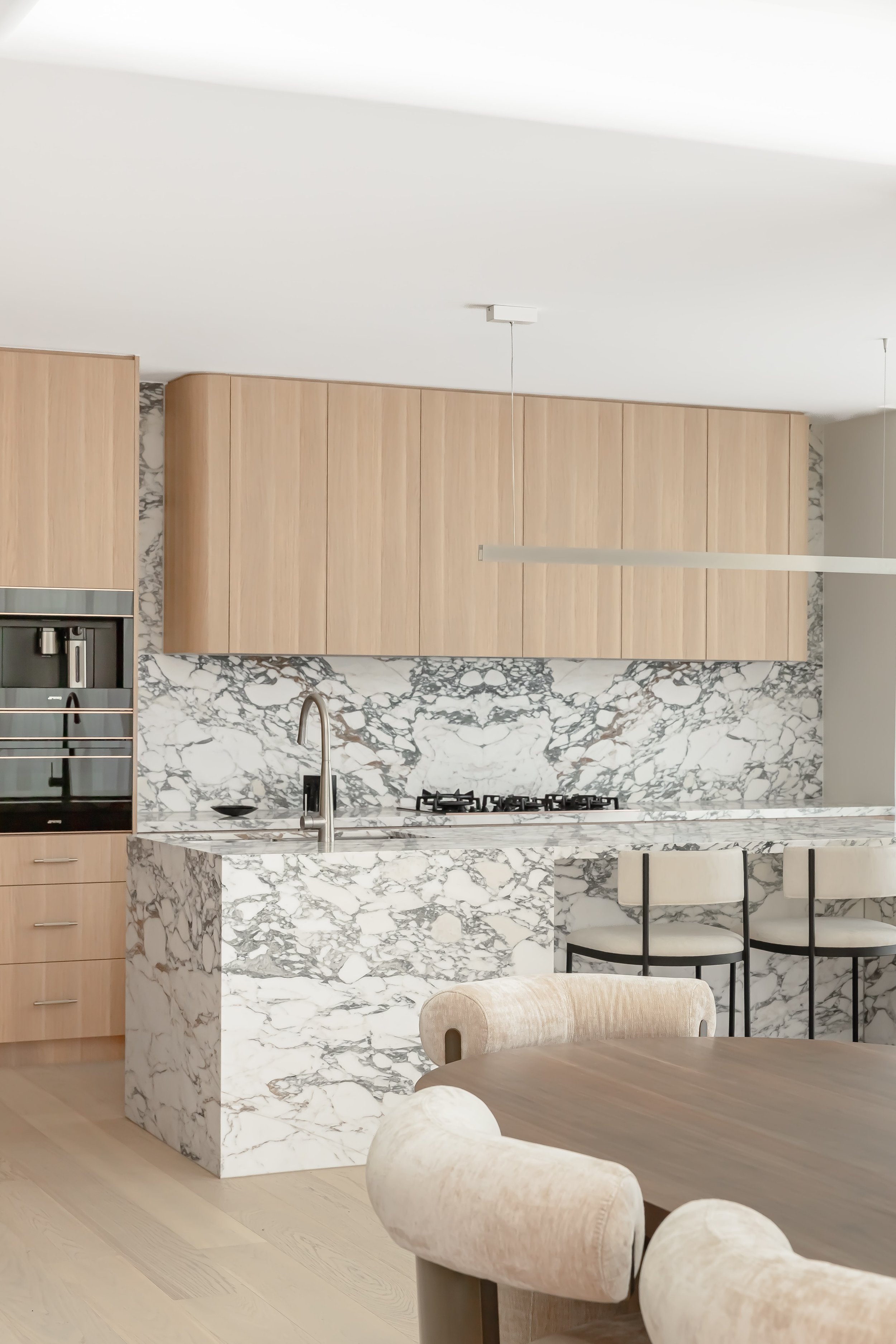 Modern kitchen with light wood cabinetry, a marble island, and marble backsplash, with a dining table and upholstered chairs in a neutral palette.